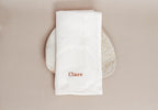 White napkin with 'Clare' embroidered on it, placed on a textured plate against a beige background.