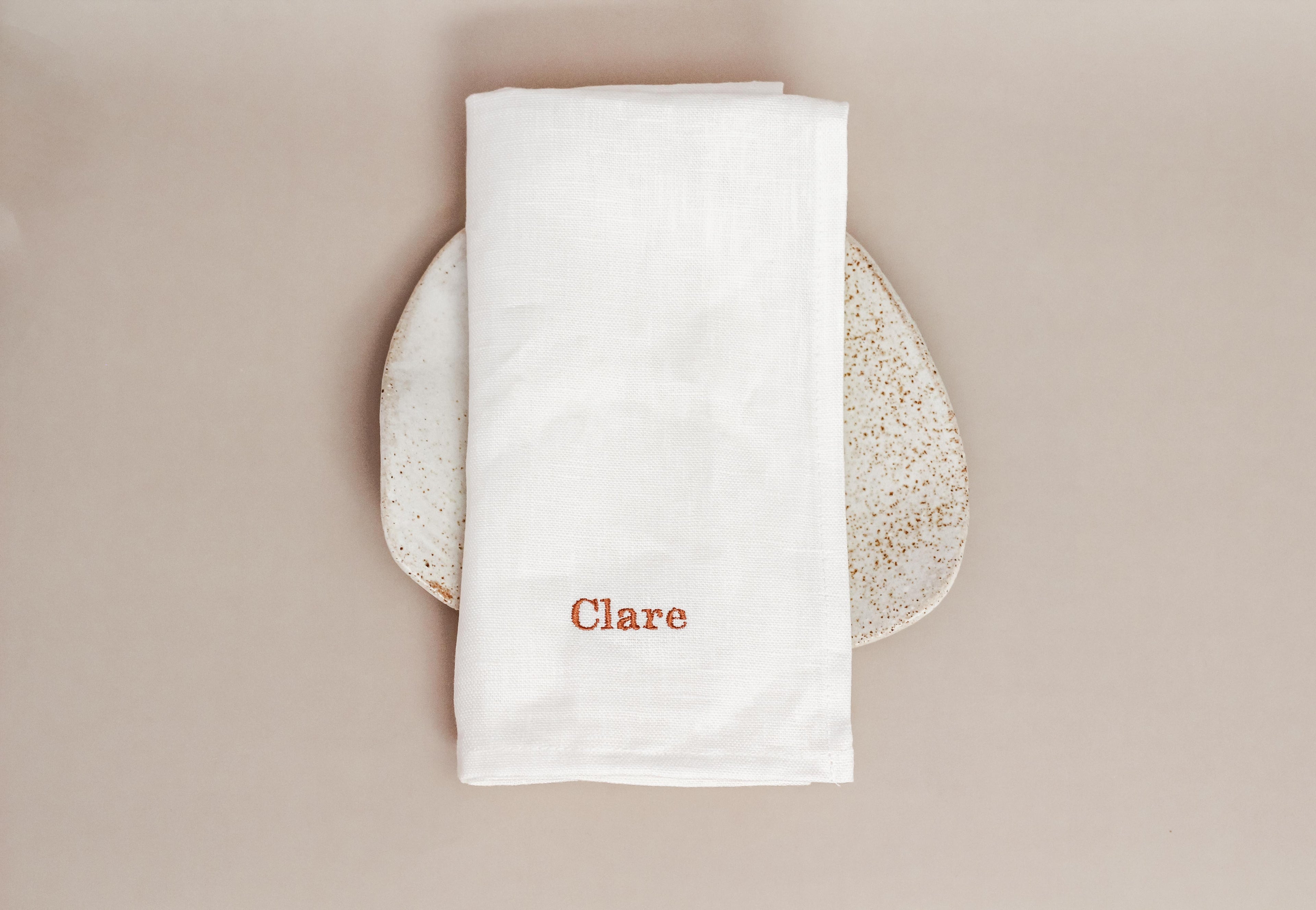 White napkin with 'Clare' embroidered on it, placed on a textured plate against a beige background.